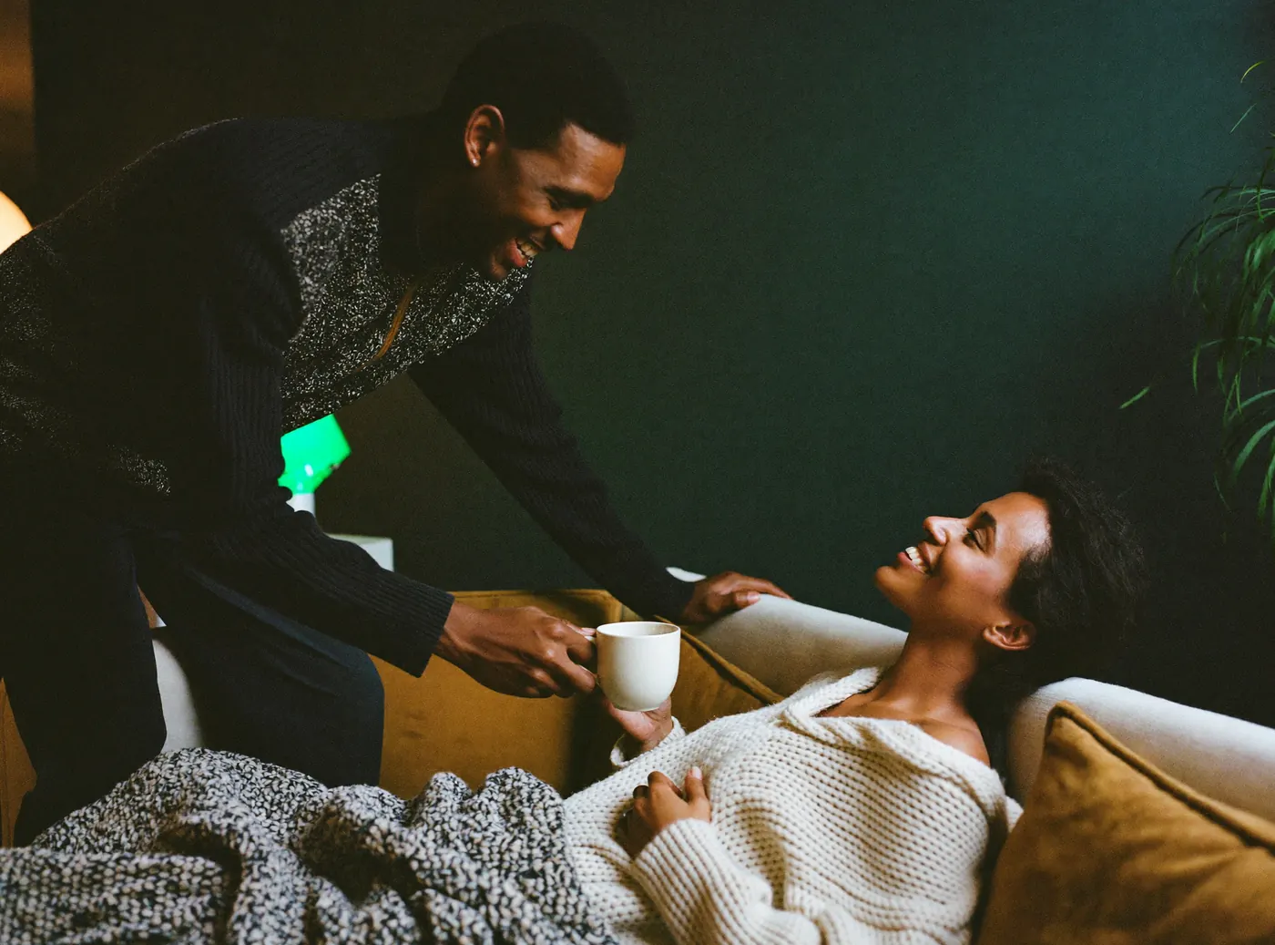 A man bringing his partner a mug of tea as she rests on the sofa under a blanket